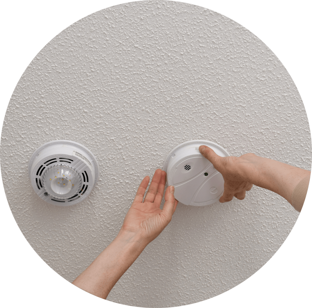 A person's hands touching a smoke alarm on the ceiling next to a carbon monoxide alarm.